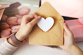 Woman's hands holding a letter in craft envelope. Pink background, valentine's day concept. Tulips flower and pink gift box in background. Womens home desk