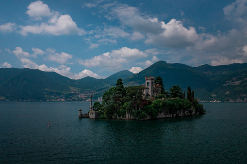 Woman stand up padling beside castle on the island Isola di Loreto on Lake Iseo, mountains in the background, during blue sky day, Italy
