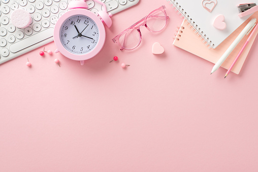 Back-to-school essentials on charming pink background: pens, stapler, notepads, clips, pushpins, glasses, hearts, keyboard, alarm clock displayed on a pastel pink backdrop. Advertise smartly