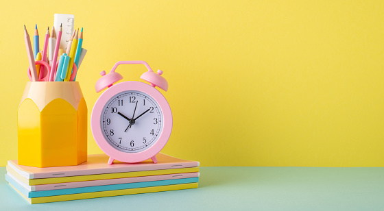 Back to school concept. Photo of school supplies on blue desktop alarm clock stack of notebooks and pencil holder on yellow wall background with empty space