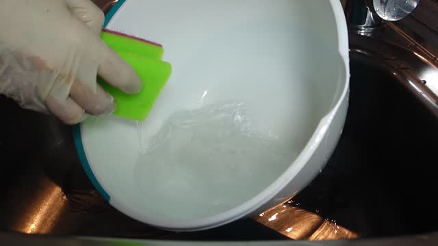 Close-up shot of a woman's hands in light gloves who wets a dishwashing sponge and washes a plastic bowl in a metal sink.
