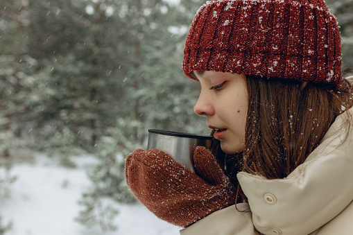 Side view of female drinking hot tea from thermos cup in snowy winter forest