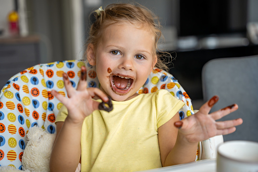 Little girl with blond hair eating homemade chocolate and showing mouth and dirty hands with stains of chocolate in home kitchen. High quality photo