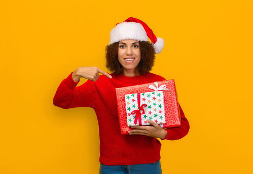 Young woman in santa claus and knitted red sweater holding gift boxes standing on yellow background, big christmas discounts, sale concept
