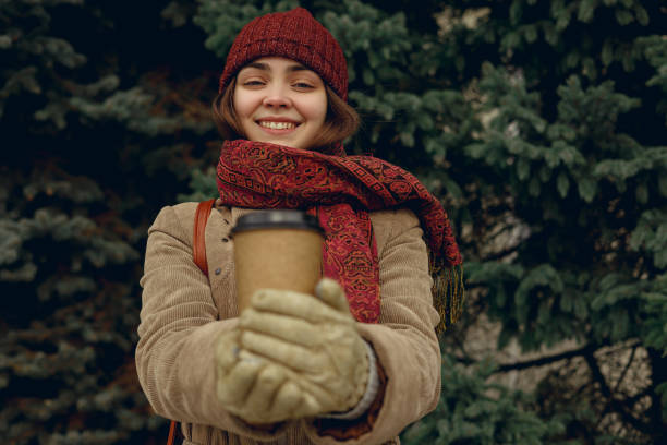 Cheerful female warming hands with coffee in takeaway blank paper cup in cold weather in winter park