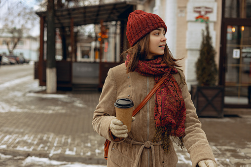 Stylish female in warm hat and coat with cup of takeaway coffee crossing road in city in winter and looking away