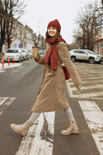 Side view of smiling female in coat walking with takeaway coffee and crossing road in autumn while looking at camera