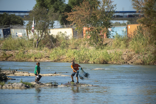 A man casts a net into the Rio Grande on Saturday, August 5, 2023 in Piedras Negras, Mexico. A barricade of shipping containers lined with razor wire can be seen on the United States side of the riverbank.