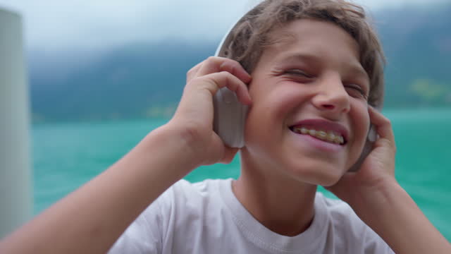 Joyful young boy listening to music wearing headphones standing by lake. Close-up face of teenager kid holding headphones over the ear bouncing head to the beat of song