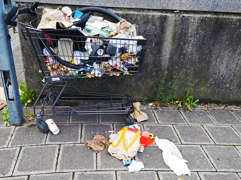 Nuremberg, Germany - shopping cart used as garbage basket crowded with paper, plastic and many widespread fast food packages on the ground.