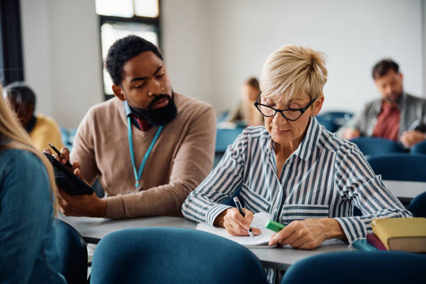 mature woman studying with help of a teacher during adult educational training class in lecture hall. - mature student learning training computer imagens e fotografias de stock