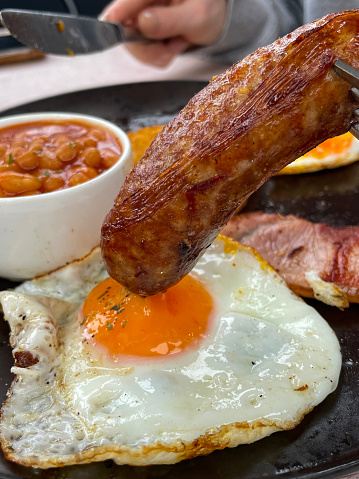Stock photo showing close-up, elevated view of a white plate containing a Full English breakfast with  ramekin of baked beans surrounded by sausage, bacon rasher, mushrooms, tomato, harsh brown, sunny side up fried egg and white toast.