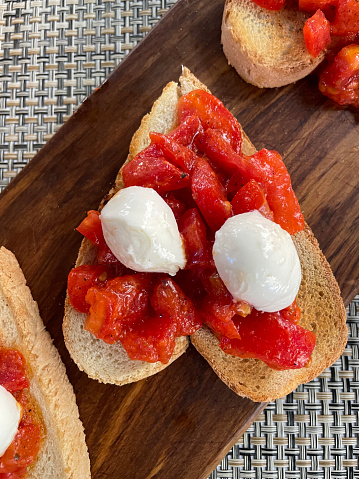 Stock photo showing close-up, elevated view of of bruschetta recipe open sandwiches with toasted, heart-shaped, white bread slices rubbed with garlic cloves and topped with diced tomatoes and buffalo mozzarella cheese ball halves, drizzled with balsamic vinegar and extra virgin olive oil on wood grain cutting board.
