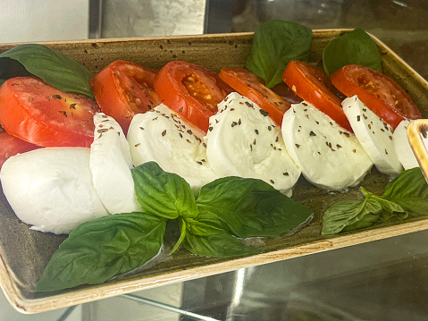 Stock photo showing close-up view of restaurant display shelf containing tray of Caprese salad with rows of beef tomato slices, slices of Buffalo mozzarella and basil leaves, available for purchasing.