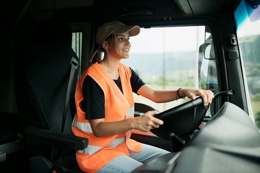 Happy young woman driver wearing cap and reflective vest driving a truck. Female trucker driving a delivery vehicle.