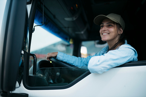 Smiling young woman driver in a truck. Caucasian female sitting on driving seat of a truck looking out the window and smiling.