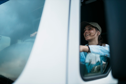 Beautiful young woman driving a semi-truck. Happy female truck driver on driving seat of truck seen through window.