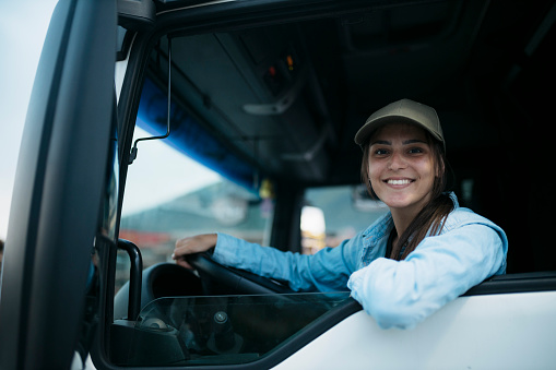 Attractive young woman sitting in drivers seat with hands on steering wheel and smiling at camera. Portrait of smiling caucasian female truck driver driving a semi-truck.