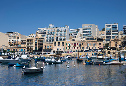 Fishing boats in Spinola Bay in the St. Julian's region of Malta with multistorey buildings in the background and tall palm trees in the foreground. The sky is clear blue with no clouds. The walkway around the bay is a quiet getaway from the bustle of the main streets.