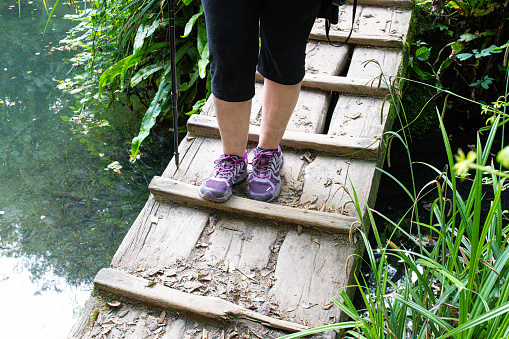 woman's legs and sneakers with a trekking pole walking over an old wooden bridge over a small river in nature