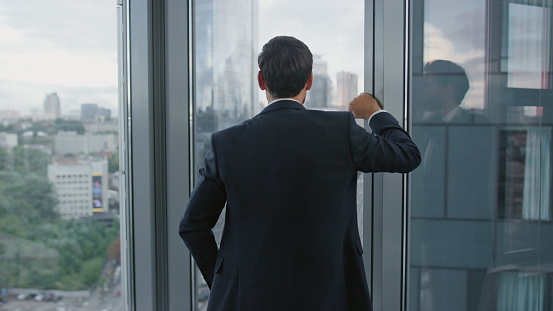 Confident director crossing hands standing near panoramic office window in elegant suit closeup. Portrait of satisfied business man thinking about own profitful company financial success. Manager work