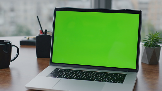 Modern green screen laptop standing on office wooden desk close up. View of chroma key computer on table with cup coffee plants pots. Turned on greenscreen mockup monitor notebook ready for work.