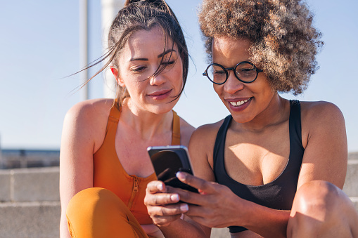 two female runner friends using a mobile phone sitting outdoors, concept of friendship and sporty lifestyle