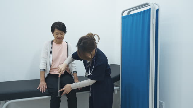 Asian chinese female doctor using reflex hammer tapping on senior woman knee at clinic consultation room checking nervous system