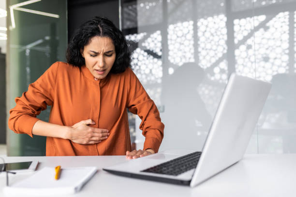 istock Sick business woman working inside office using laptop while sitting at table, Hispanic woman has severe stomach pain 1599879152