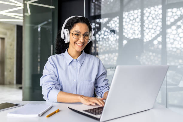 istock Young beautiful Indian female programmer working inside office with laptop, woman in headphones listening to audio podcast and online course, typing on keyboard and reaching out, female engineer 1599879171