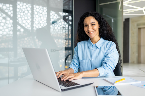 Young beautiful Indian female programmer working inside the office on a laptop, woman smiling and looking at the camera, portrait of a satisfied and confident businesswoman at the workplace.