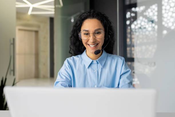 istock Young beautiful woman with video call headset working inside office at workplace, Hispanic online customer support worker typing on keyboard looking at laptop screen 1599879276