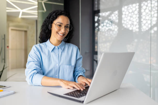 istock Young beautiful woman with video call headset working inside office at workplace, Hispanic online customer support worker typing on keyboard looking at laptop screen 1599879280