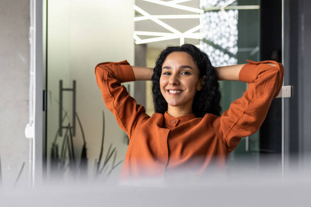 satisfaite d’elle-même et du travail accompli, une jeune femme d’affaires latino-américaine est assise dans le bureau sur une chaise et se repose, les mains derrière la tête, souriante - yoga business women indoors photos et images de collection