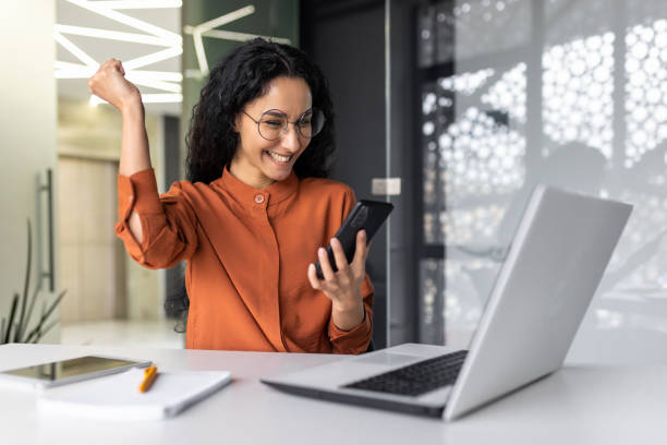 istock Young successful hispanic businesswoman working inside modern office, female worker using laptop, holding phone smiling and happy celebrating victory and triumph 1599879748