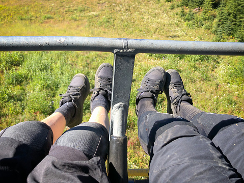 Personal perspective of senior Chinese mother and Eurasian adult daughter mountain bikers wearing knee pads and biking gear riding chairlift with alpine meadow and forest below.