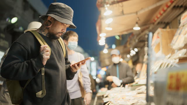 Asian man is visiting the night market in the evening looking for dry food and use mobile for calculation price.