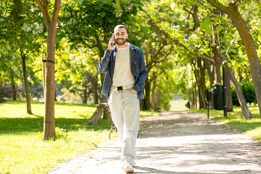 A young caucasian man, engaged in a phone conversation, takes a leisurely walk along a lush green path surrounded by abundant vegetation