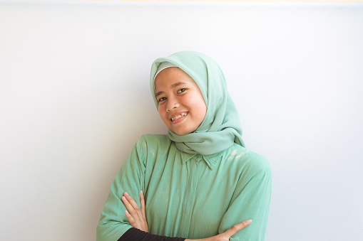 Portrait of an Asian Muslim girl smiling with toothy smile with arms crossed looking at the camera with a white background, she's wearing muslim dress, natural expression