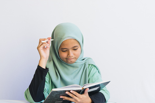 A serious young Asian Muslim girl uses a pen on her head to think while holding a notebook on a white background, a medium shot and space for copy