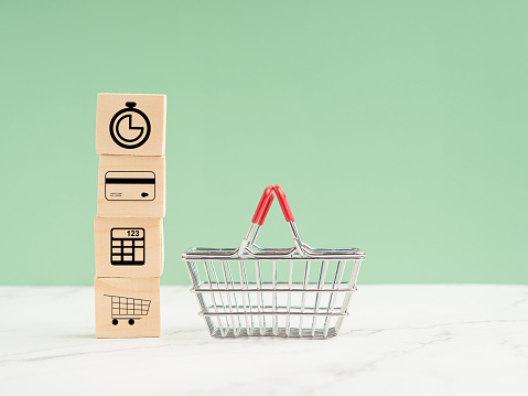 Business shopping online concept. Wooden cubes with icons of the time, a credit card, a calculator, a shopping trolley. A shopping basket over a marble floor against a light green background