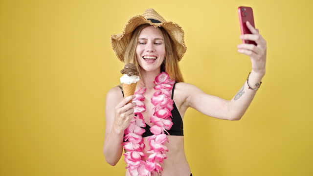 Young blonde woman tourist holding ice cream make selfie by smartphone over isolated yellow background