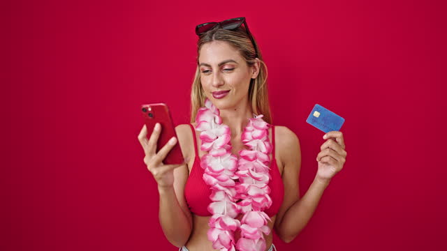 Young blonde woman wearing bikini shopping with smartphone and credit card over isolated red background