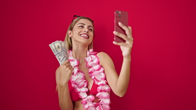 Young blonde woman wearing bikini holding dollars having video call over isolated red background