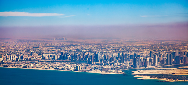 Foggy Dubai, United Arab Emirates, with skyscrapers and high-rise buildings densely packed along the coastline. The city has a sandy beach and a harbor with boats, and the sea is calm and blue. The sky is clear with a few clouds and a pinkish hue on the horizon. The photo is taken from a high vantage point, looking down on the cityscape.