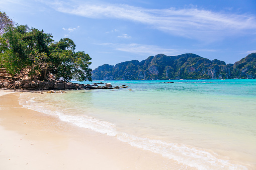 A tropical beach in Phi Phi Don Island, Thailand, with clear blue water and white sand. The beach is surrounded by green trees and rocks on one side and a mountain range on the other side. The water is a beautiful shade of turquoise and is very clear, reflecting the sky and the mountains. The sky is blue with a few clouds, creating a contrast with the green landscape. The sand is white and there are a few rocks scattered around, adding some texture to the image. The overall mood of the image is peaceful and serene, inviting the viewer to relax and enjoy the scenery.