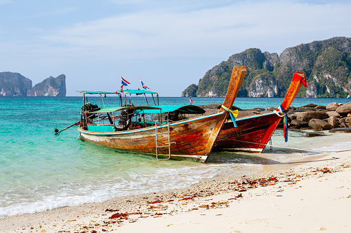 Two wooden boats on a sandy beach with clear blue water and rocky cliffs in the background. The boats are traditional Thai longtail boats with colorful decorations on the bow. The boats are moored on the beach with ropes and anchors. The water is clear and turquoise in color. The cliffs in the background are tall and rocky with green vegetation on top. The sky is blue with a few clouds. The image shows a typical scene on Koh Phi Phi Don island, a popular tourist destination in Thailand.\n4
