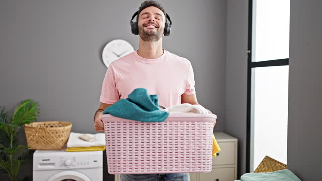 Young hispanic man listening to music holding basket with clothes dancing at laundry room