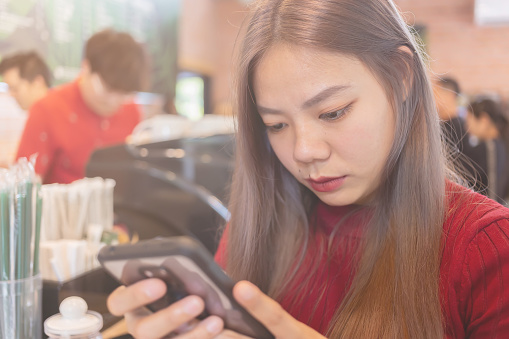 Asian woman is using a smartphone in a coffee shop with white sceen and clipping path. Mobile phones with white screens on the hands of women in convenience stores.