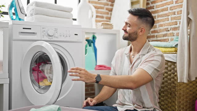 Young hispanic man washing clothes holding dirty t shirt at laundry room